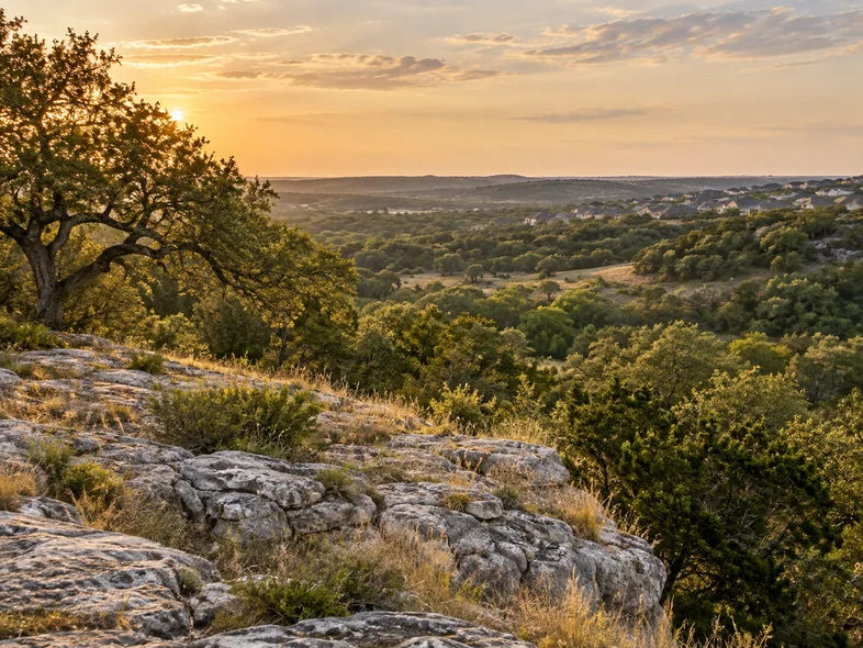 Rolling Hill Country landscape with live oaks, limestone outcrops and distant rooftops at golden hour